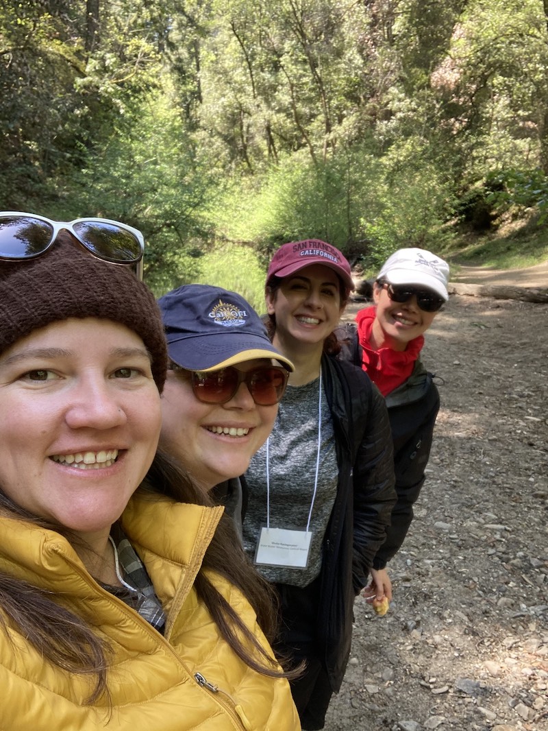 photo of four women smiling, standing on a path in a forest