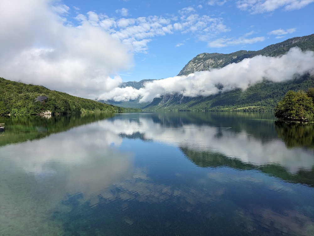 Photo of Lake Bohinj in Slovenia. Green hills to left and right of lake. Blue sky, dotted clouds and some low strips of cloud or mist reflected in the lake surface