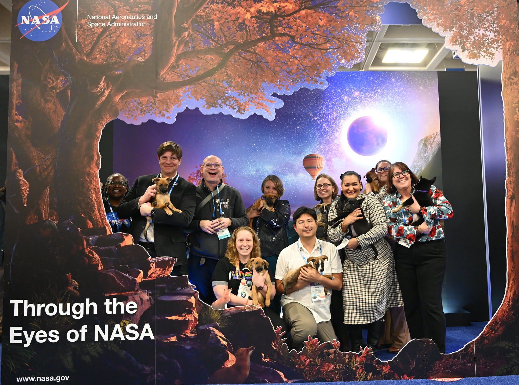 10 people standing or kneeling, holding puppies and smiling, against a backdrop that says 'through the eyes of NASA' with nasa logo and tall trees and night sky trees