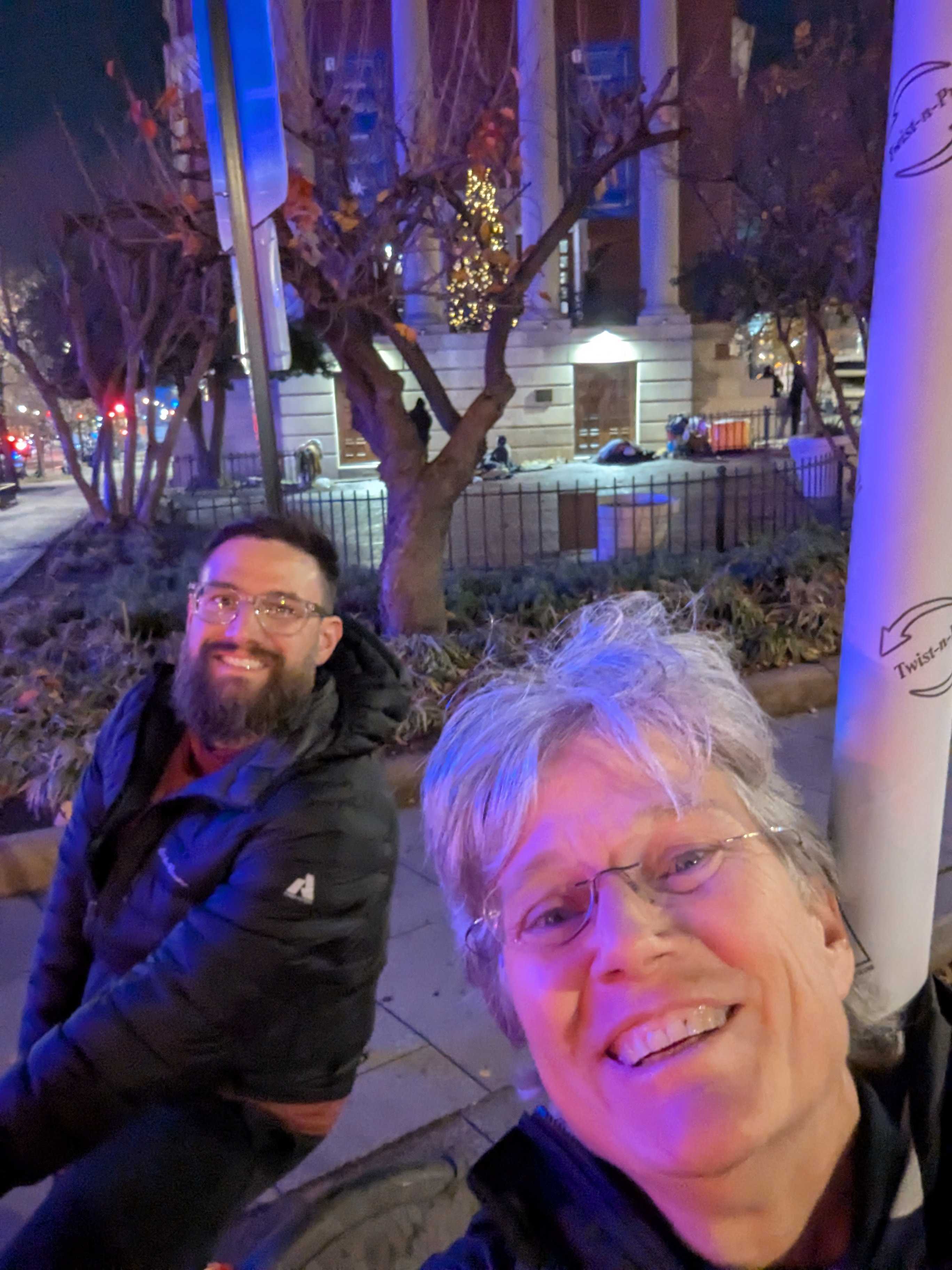 selfie of a woman and man, sitting on bikes that are out of view, in front of a white building and a tree and street signs. Dark outside. Colored lights in the background.