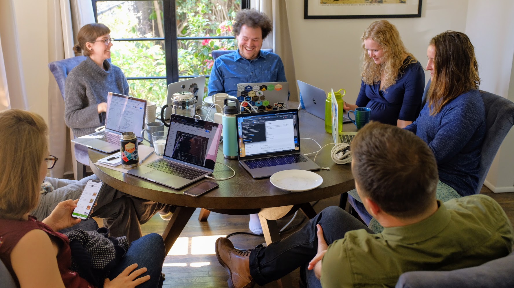 6 smiling people sit at a round table with their laptops open. Window in background