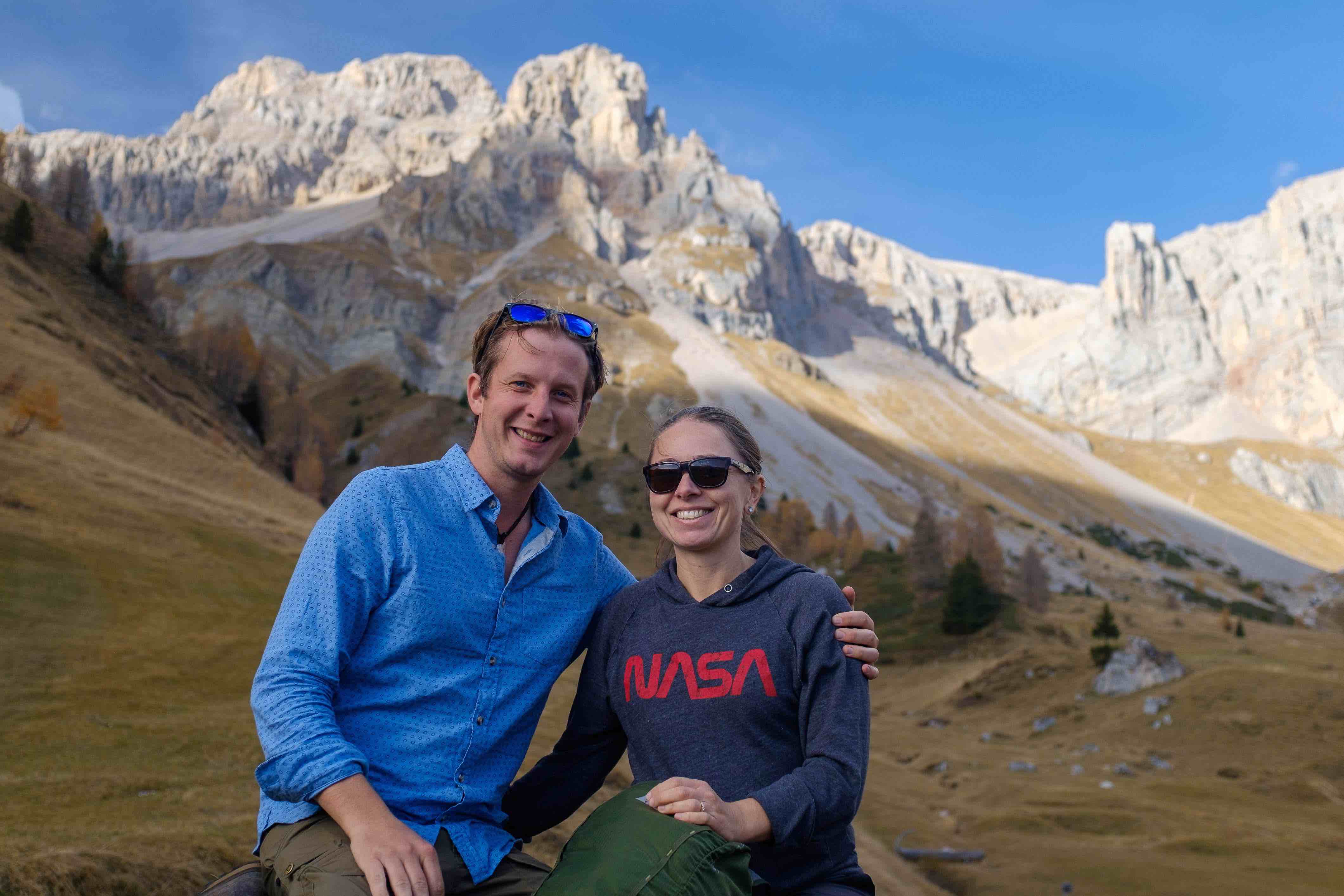 photo of a white man and woman smiling, wearing hiking clothes with mountain peaks behind them